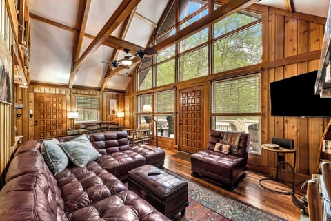 The main living room features soaring ceilings, a wall of windows with forest views, a 50" TV, and a luxurious leather sectional—perfect for cozy nights, movie marathons, or simply soaking in the rustic charm.