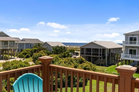 Upper deck view with wooden railing, turquoise chair, neighboring beach homes, lush greenery, and a blue ocean peek near Oak Island's shore.