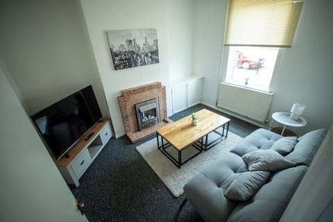 Cozy living room with a grey sofa, wooden coffee table, stylish fireplace, and flat-screen TV. Bright space with natural light from the window, modern decor, and neutral tones create a warm, relaxing atmosphere.