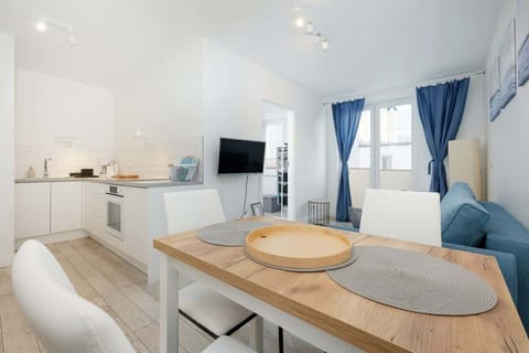 A modern living room with a wall-mounted TV, blue sofa, white furnishings, and ample natural light.
