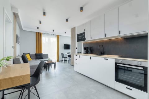 The modern, open space of the kitchenette and living room impresses with sleek white cabinets with black handles, a stylish black panel over the countertop, and a comfortable gray sofa with decorative pillows in shades of yellow and gray, creating a