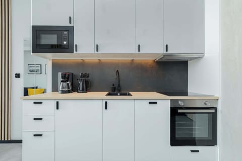 A stylish kitchenette with white cabinets, black countertop and dark gray wall panel, equipped with modern appliances - microwave, oven and coffee maker - and under-cabinet lighting that adds elegance and functionality to the interior.