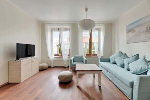 : A cozy living room featuring a pair of soft turquoise sofas and a circular wooden coffee table. Large windows fill the space with warm sunlight, making it an inviting spot for relaxation.