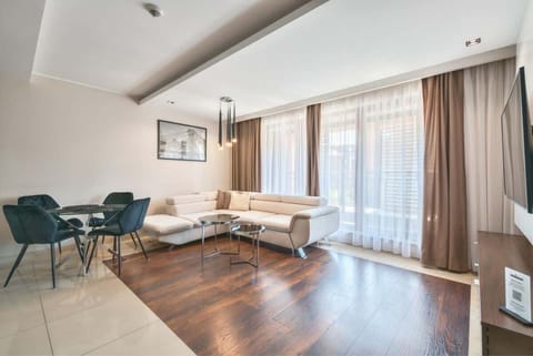 A spacious living room with a modern design, featuring a wooden floor, a glass coffee table, and comfortable seating. Large windows provide plenty of natural light.