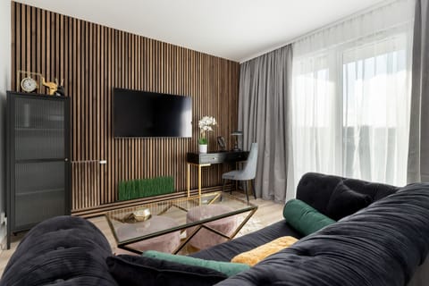 A bright living room  with a TV, a comfortable seating arrangement, and large windows with sheer curtains. The open layout combines modern design with a cozy feel.