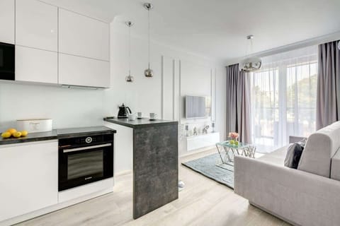 A modern open-concept living area featuring a sleek white kitchen with a gray breakfast bar, a cozy living space with a light gray sofa, a glass coffee table, and large windows with sheer curtains that fill the room with natural light.