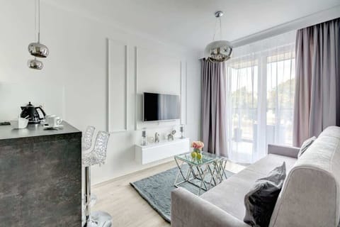 An elegant living room with a light gray sofa, a glass coffee table, decorative wall paneling, a mounted TV, and floor-to-ceiling windows with sheer and heavy curtains, creating a bright yet sophisticated atmosphere.