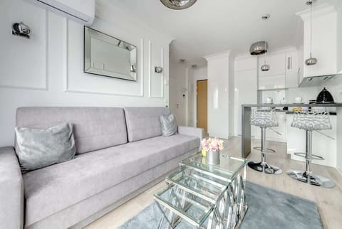 A modern open-plan living area with a sleek gray sofa, a mirrored glass coffee table, a minimalist kitchen with a breakfast bar and transparent bar stools, and elegant decorative accents like a wall mirror and pendant lights.