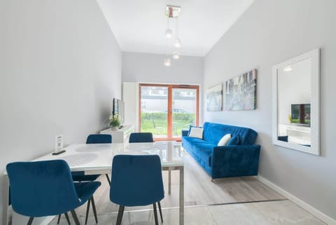 The living room features a modern design with a blue velvet sofa, a dining table with matching chairs, and large windows overlooking a green outdoor space, creating a bright and elegant ambiance.