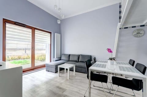The room features a modern design with a gray sofa, a sleek dining table with black chairs, and large windows providing natural light, complemented by a bright orchid as a decorative touch.