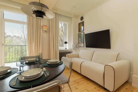 A cozy living room with a light sofa, round dining table, and TV, featuring large windows letting in natural light.