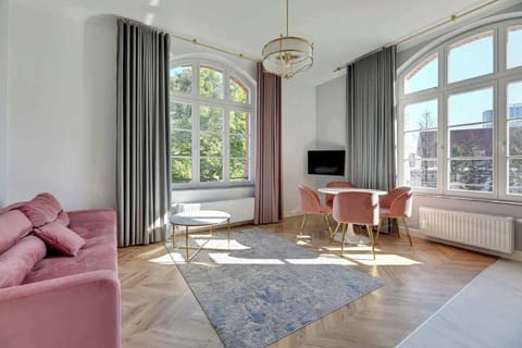 Chic, elegant living space is bathed in natural light, with soft pink velvet furnishings and sophisticated gold accents, blending contemporary style with timeless charm in a bright, airy atmosphere.