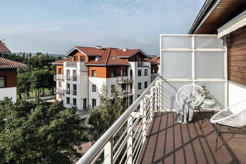 A spacious outdoor balcony with a set of chairs and a small round table. The view overlooks red-roofed buildings and a beautiful, sunny neighborhood.