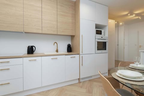 The modern kitchenette impresses with an elegant combination of light wood and white, gold cabinet handles, integrated appliances and a stylish dining table with a cover, creating a functional and aesthetically pleasing space for cooking and dining.