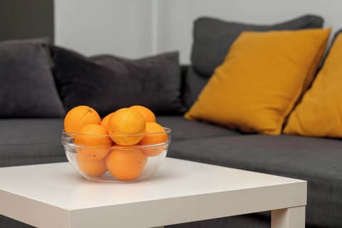 A close-up of a white coffee table with fresh oranges and a dark gray sofa with yellow pillows in the background.