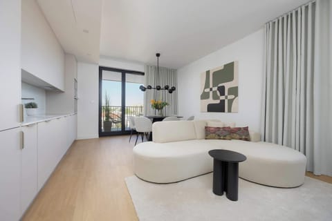 A bright open-plan room with a white sofa, light wood floors, and a small dining nook near the window.