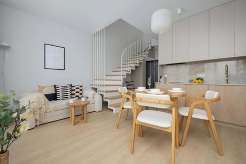 A bright and elegant living room with a dining area and light wooden flooring. The space features modern lighting and minimalistic décor.