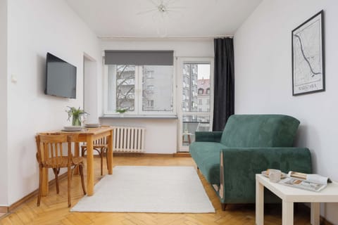 A bright living room with a green armchair, a wooden dining table, and a TV mounted on the wall. The open space connects to the kitchen.