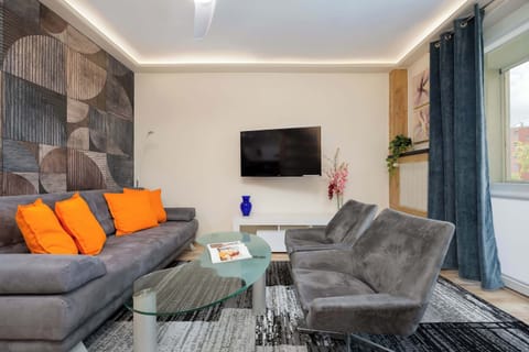 A modern living room with a gray sofa and two bright orange armchairs, a mounted TV, and a wood-accent wall.