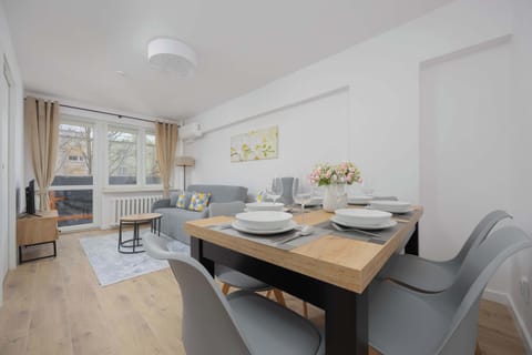 A cozy living room with a dining table for four, soft-toned decor, and natural light streaming through the windows.
