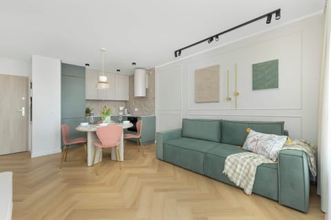 Modern and cozy living room with a green sofa, wooden floor and bright walls. In the background you can see a section of the kitchenette with pink accents.