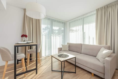 A cozy living room with a light-colored sofa placed next to a window, which allows plenty of natural light. A simple round coffee table sits in front of the sofa, and a large houseplant adds greenery to the space.
