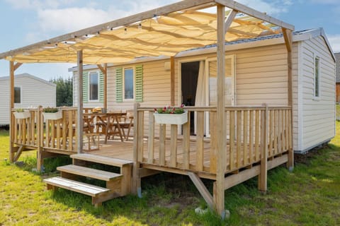 A cozy mobile home with a spacious wooden terrace covered by a roof, surrounded by a green field and scenic countryside.