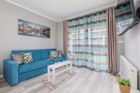 A cozy living room with a blue sofa, a coffee table, and a window with colorful curtains. The space is well-lit and decorated with paintings on the wall.