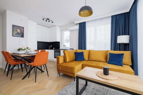 A bright living room with a comfortable mustard-yellow sofa, coffee table, and a TV stand. The room is filled with natural light from large windows and decorated with modern touches.