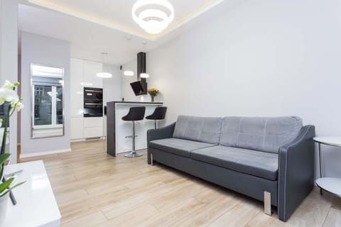A modern living room with a gray sofa, a white TV stand, and a glass door leading to a balcony. The space is illuminated by natural light.