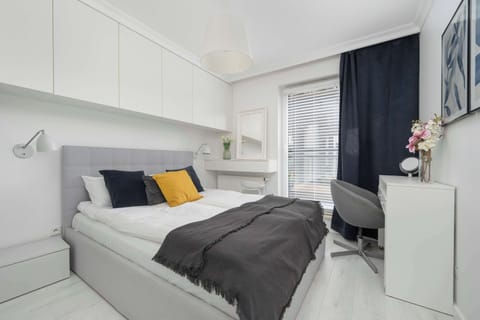 A bright bedroom featuring a gray bed with yellow and white cushions, a small desk with a chair, and floral artwork on the wall.