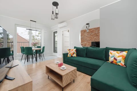 The living space features a bold brick accent wall, a green sofa, and a wooden coffee table. Decor includes colorful throw pillows and a modern TV setup.
