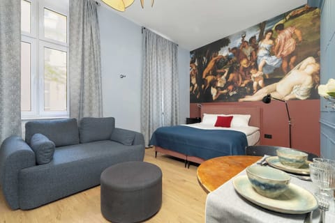 A bright living space featuring a blue sofa, round coffee tables, and a double bed with a striking classical art mural above the headboard.