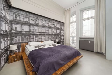A cozy bedroom with a large bed against a stylish black-and-white cityscape wallpaper. Natural light floods in through the large windows.