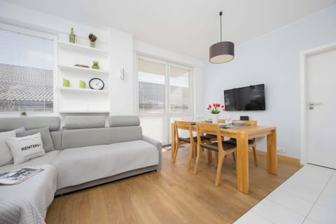 A bright living room with a gray sofa, a wooden table with two chairs, and a wall-mounted TV.