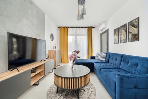 A cozy living room with a large blue sectional sofa, a wooden coffee table, a flat-screen TV mounted on the wall, and floor-to-ceiling windows allowing ample natural light to brighten the space.