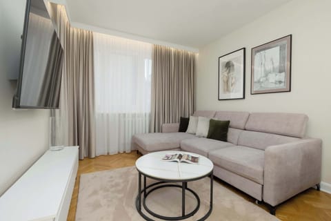 A bright living room with a gray sofa, a round coffee table, a TV on the wall, and large windows with curtains.