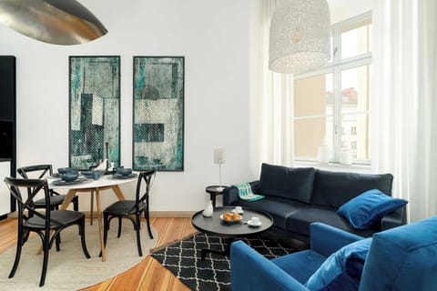 A bright living area featuring a blue sofa, a modern black coffee table, and large windows that flood the space with natural light. The wooden floors add warmth, while a patterned accent wall provides visual interest.