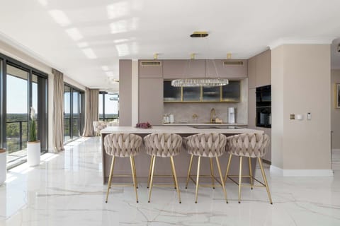 A luxurious kitchenette with an elegant island finished with a marble countertop, surrounded by four stylish hockers with beige quilted upholstery and gold legs, impresses with modern built-in appliances, a striking chandelier and large windows that 