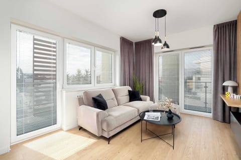 A bright and modern living room with large windows, a comfortable beige sofa, and a sleek TV mounted on a wooden accent wall.