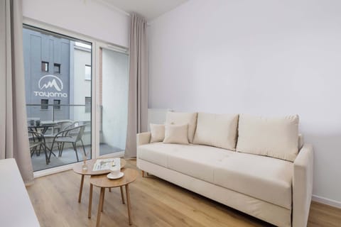 A cozy living room with a light beige sofa, a small wooden coffee table, and a large window that brings in natural light. The room has a minimalist, modern design.