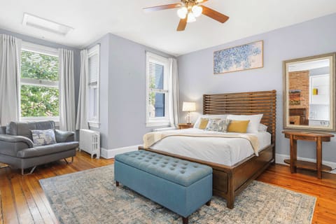 Primary bedroom with wooden floors, queen sized bed, ceiling fan, sofa, windows and a view of the outside.