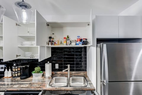 Fully equipped kitchen with double sinks, modern black subway tile backsplash, and ample white cabinet storage for all your cooking needs. Features a large stainless steel refrigerator, coffee maker, and essential spices. Perfect for home-cooked meals!