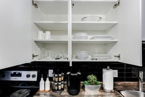 Well-stocked kitchen with open white cabinets displaying plates, glasses, and bowls. Features a coffee maker, spice rack, and stove with an oven. Enjoy cooking with essential tools and ample dishware. Perfect for home-cooked meals!