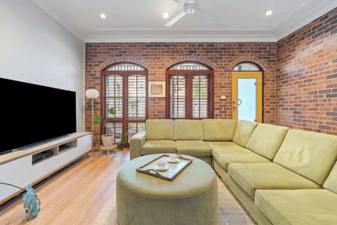 A charming lounge with a classic brick and white wall backdrop, complete with a cosy fireplace, TV, and warm sunlight streaming through.