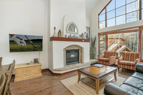 Living room with a fireplace, TV displaying a golf scene, large window offering a view of trees and a neighboring house, and a mix of seating furniture including a leather sofa and striped armchairs.