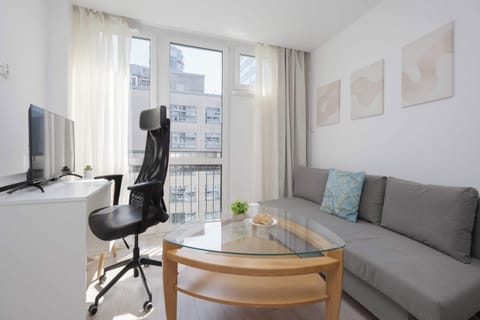 A compact living room features a grey sofa accented by a light blue pillow and three abstract wall art prints. A dedicated workspace with a white desk, an adjustable black office chair, and a computer monitor stands near the balcony window.