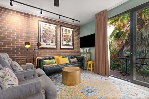 Relax in the cosy living area with a chesterfield sofa, velvet armchairs, floral art, exposed brick, soft lighting, and a wall-mounted TV—perfect for unwinding in style.