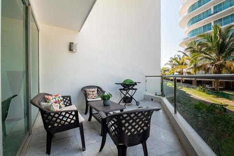 A spacious, shaded terrace equipped with comfortable wicker seating, colorful pillows, and a small grill, overlooking the resort's lush palm trees and pathways.