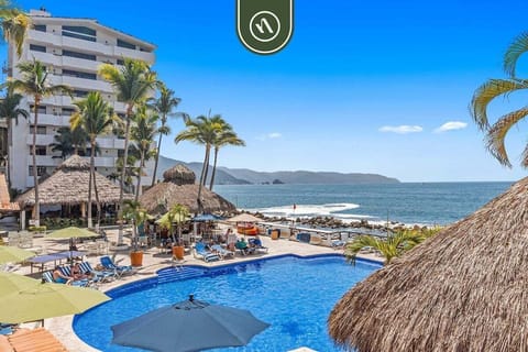 An elevated view of the beachfront swimming pool area, surrounded by thatched palapas, sun loungers, and tall palm trees against an ocean backdrop.